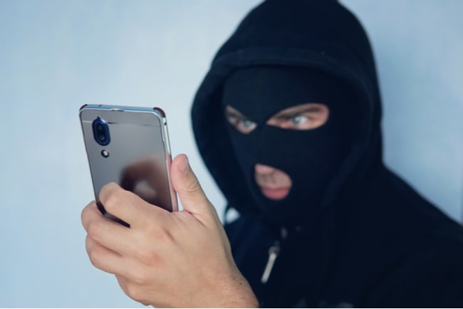 Man med svart rånarluva och svart hoodie tittar på mobiltelefon som han håller upp framför sig.