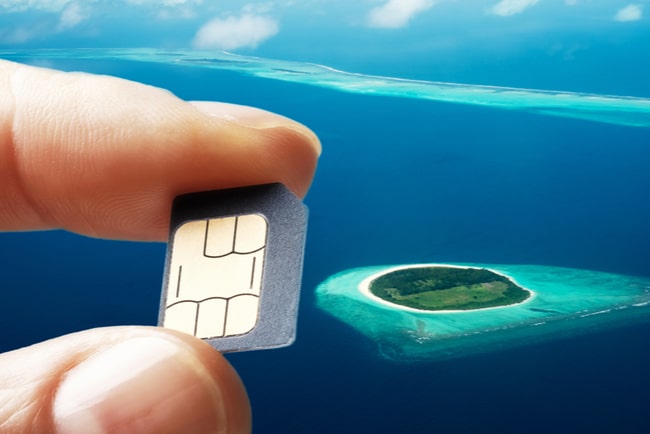 Pekfinger och tumme somhåller i ett simkort med en exotisk ö i bakgrunden.